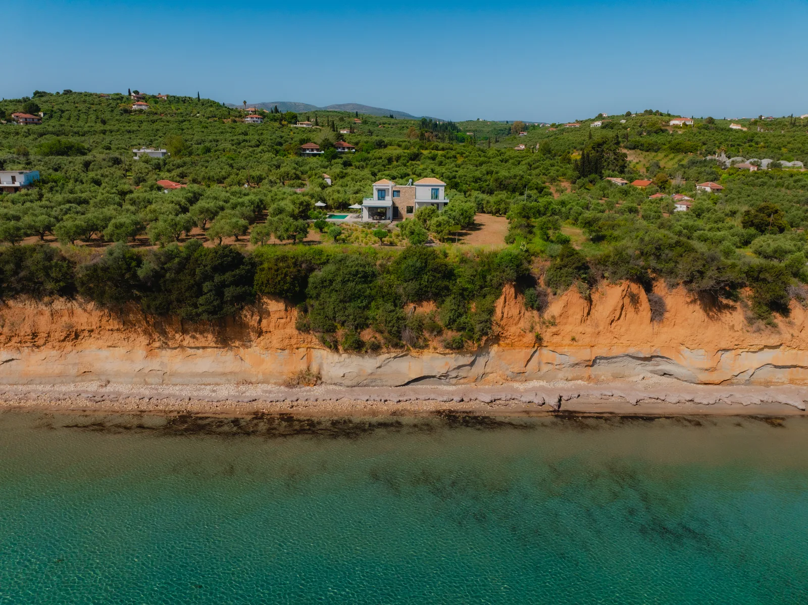 Aerial view of a house above a low cliff with olive trees and turquoise sea